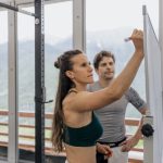 Female fitness coach writes on whiteboard as a male athlete watches in a gym.
