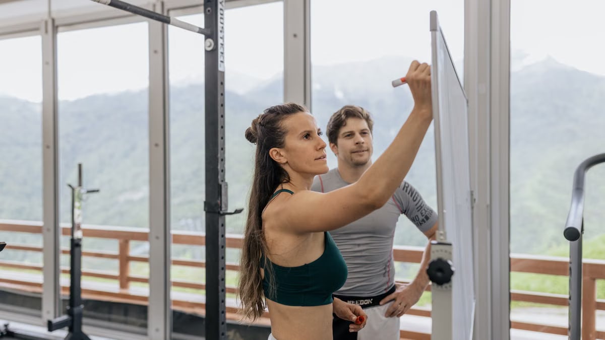 Female fitness coach writes on whiteboard as a male athlete watches in a gym.